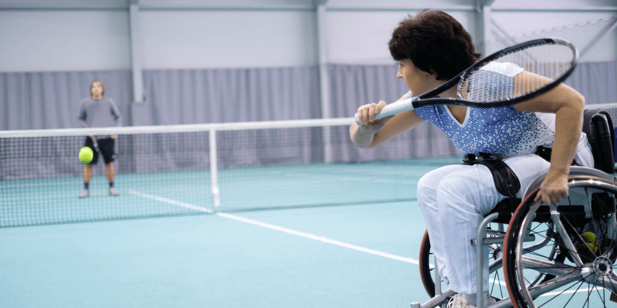 Woman in a wheelchair playing tennis against a standing opponent in a gymnasium