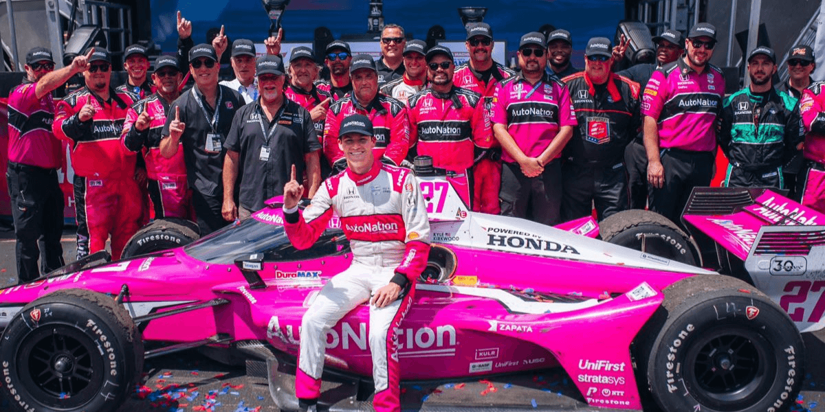 Racing team celebrates with driver next to a pink race car after a victory