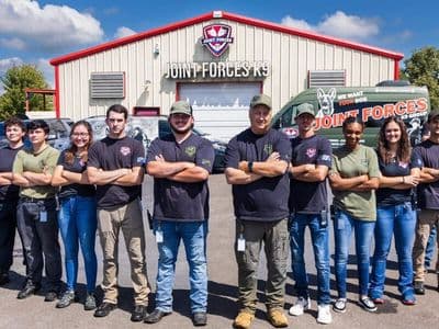 Joint Forces K9 Group team standing outside its training facility with branded vehicles behind them.
