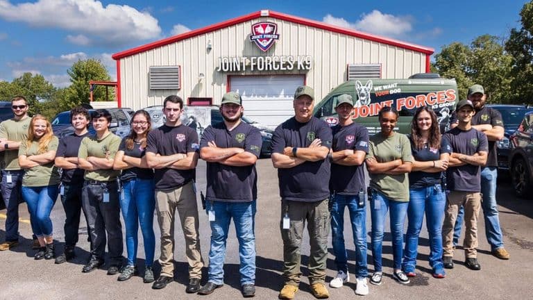 Joint Forces K9 Group team standing outside its training facility with branded vehicles behind them.