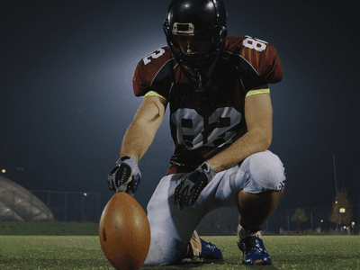 Football player kneels on a field holding a football before play under stadium lights.