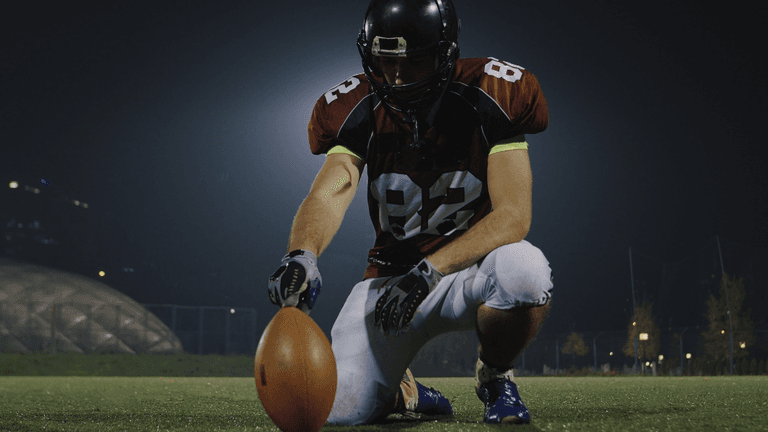 Football player kneels on a field holding a football before play under stadium lights.