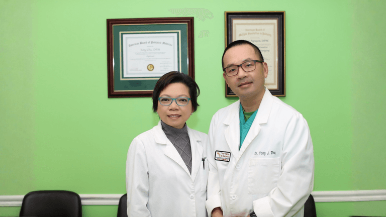 Two podiatrists in white coats standing in clinic office with framed certificates on green wall.