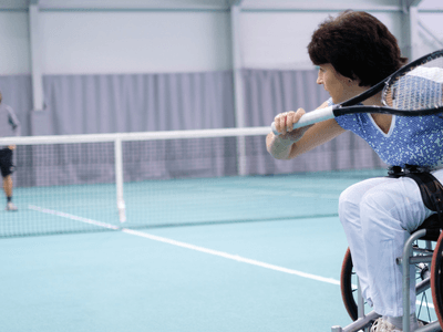 Woman in a wheelchair playing tennis against a standing opponent in a gymnasium