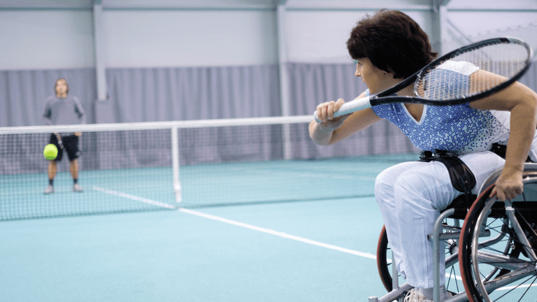 Woman in a wheelchair playing tennis against a standing opponent in a gymnasium