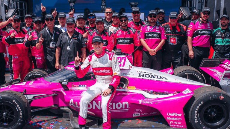 Racing team celebrates with driver next to a pink race car after a victory