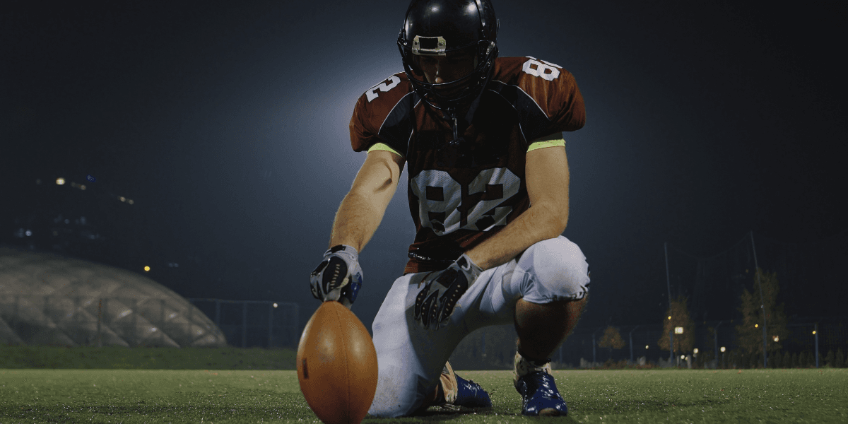 Football player kneels on a field holding a football before play under stadium lights.